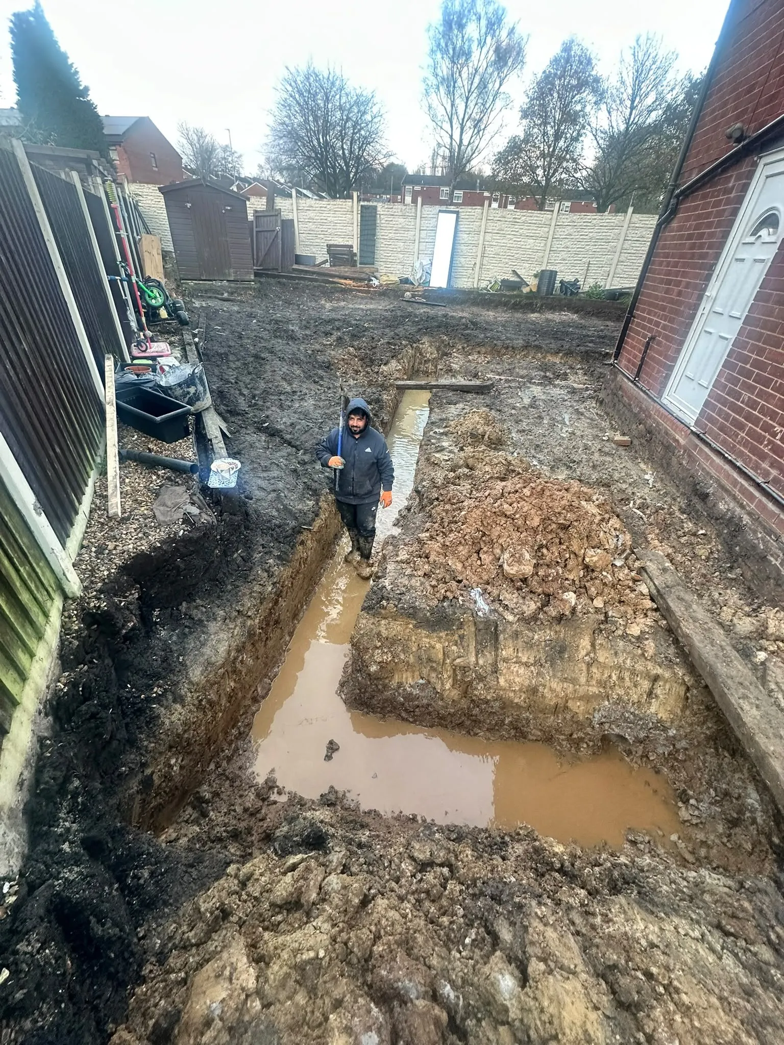 Worker standing in a deep foundation trench dug behind a house